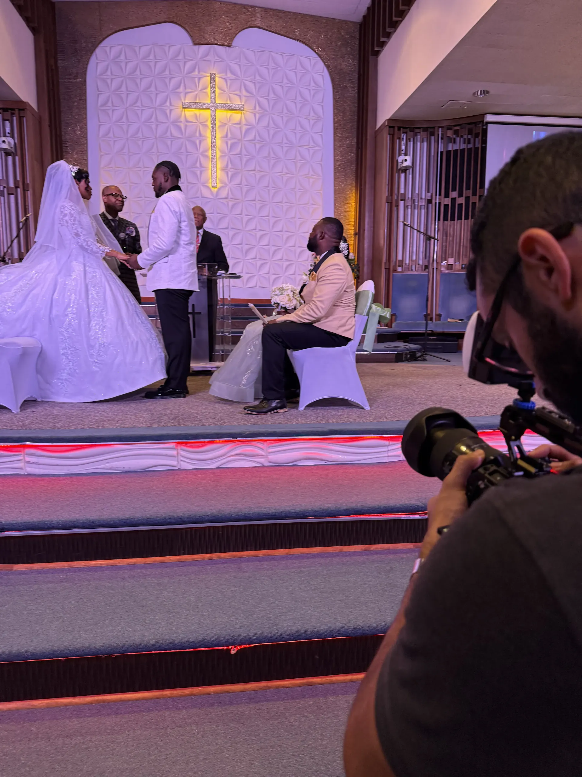 A wedding ceremony takes place in a church, seen from behind a photographer holding a professional camera.