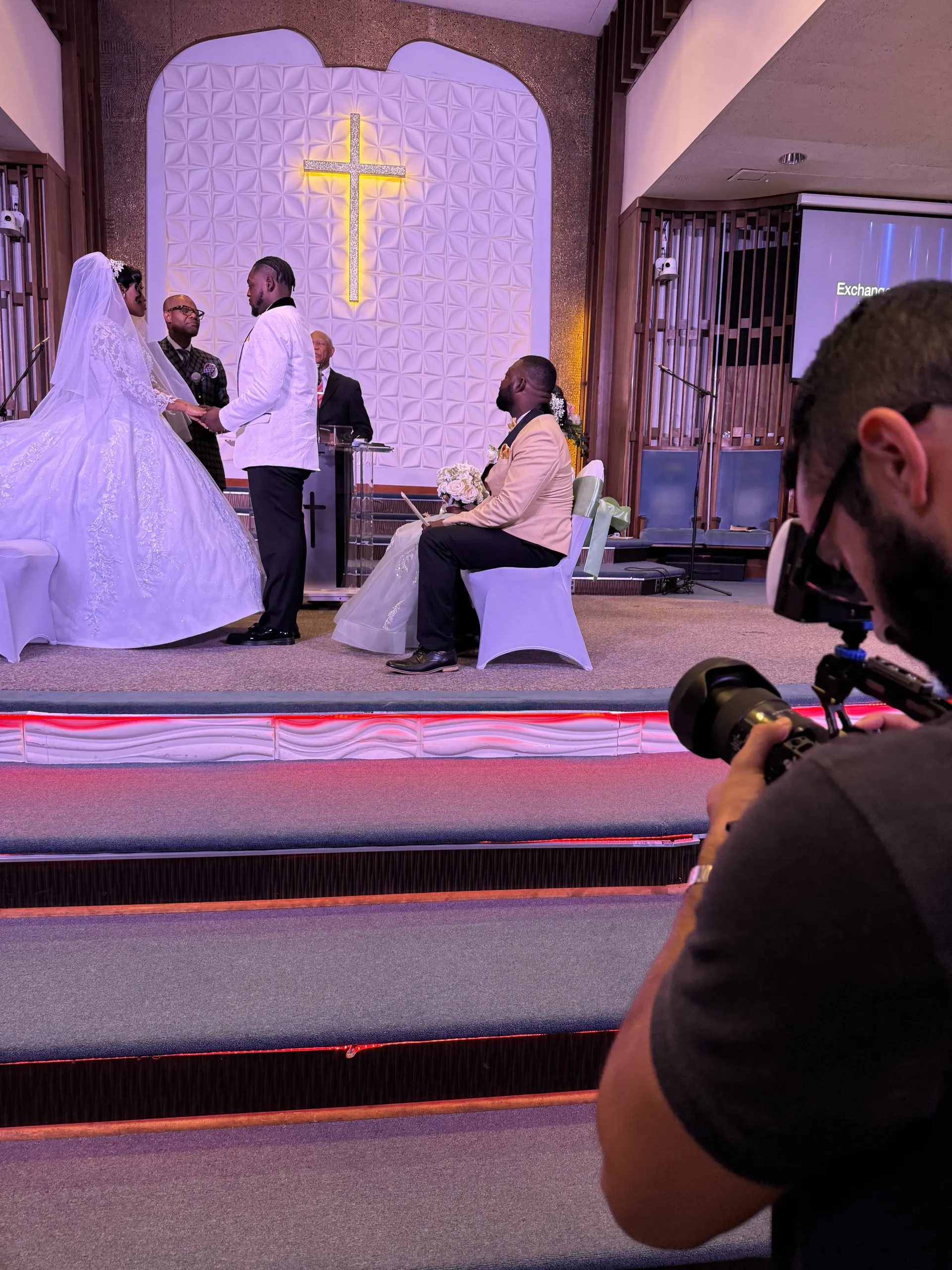 A photographer captures a wedding ceremony at an altar with a large glowing cross in a church.