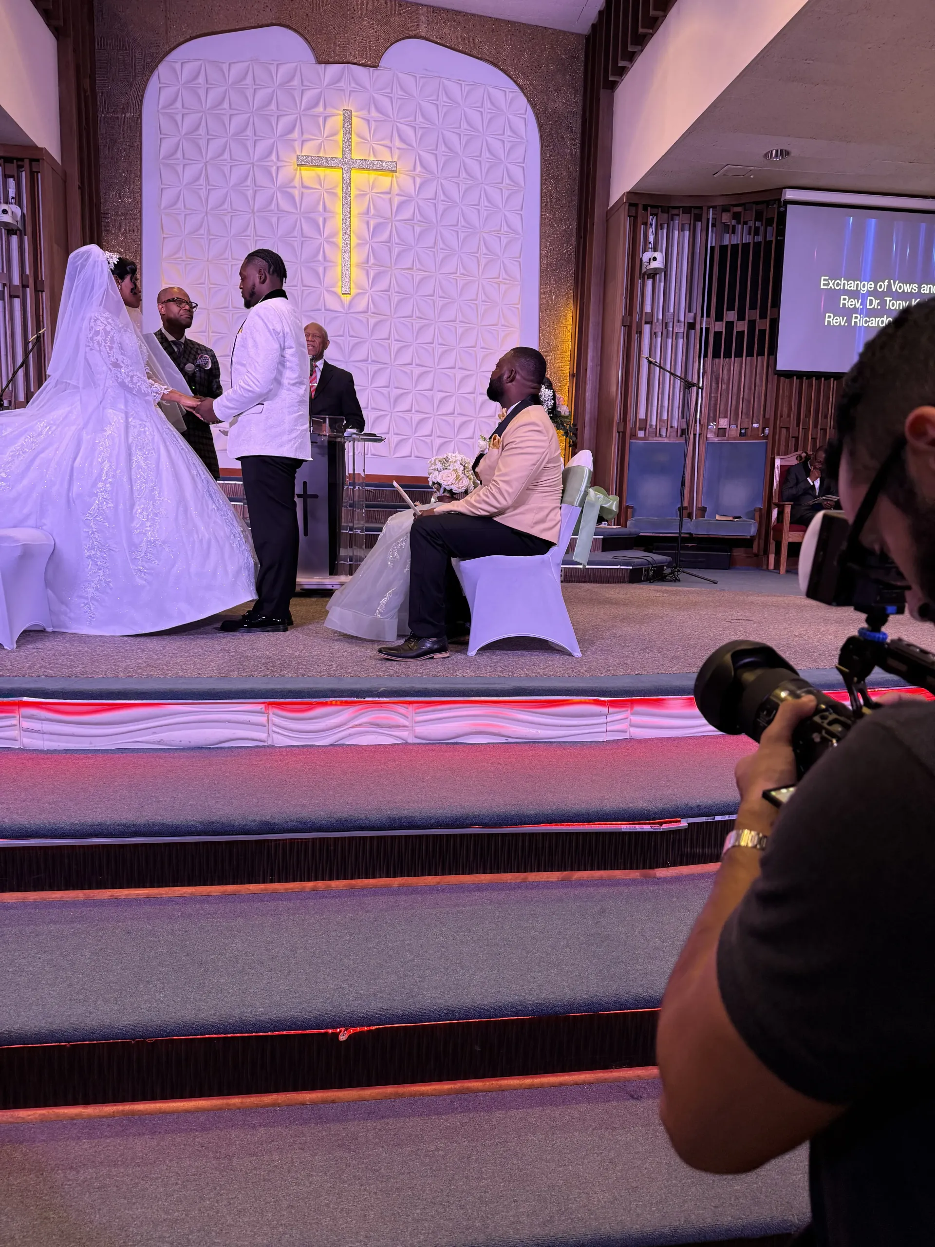 A videographer captures a wedding ceremony in a church with a gold cross backdrop, featuring a bride and groom at the altar.