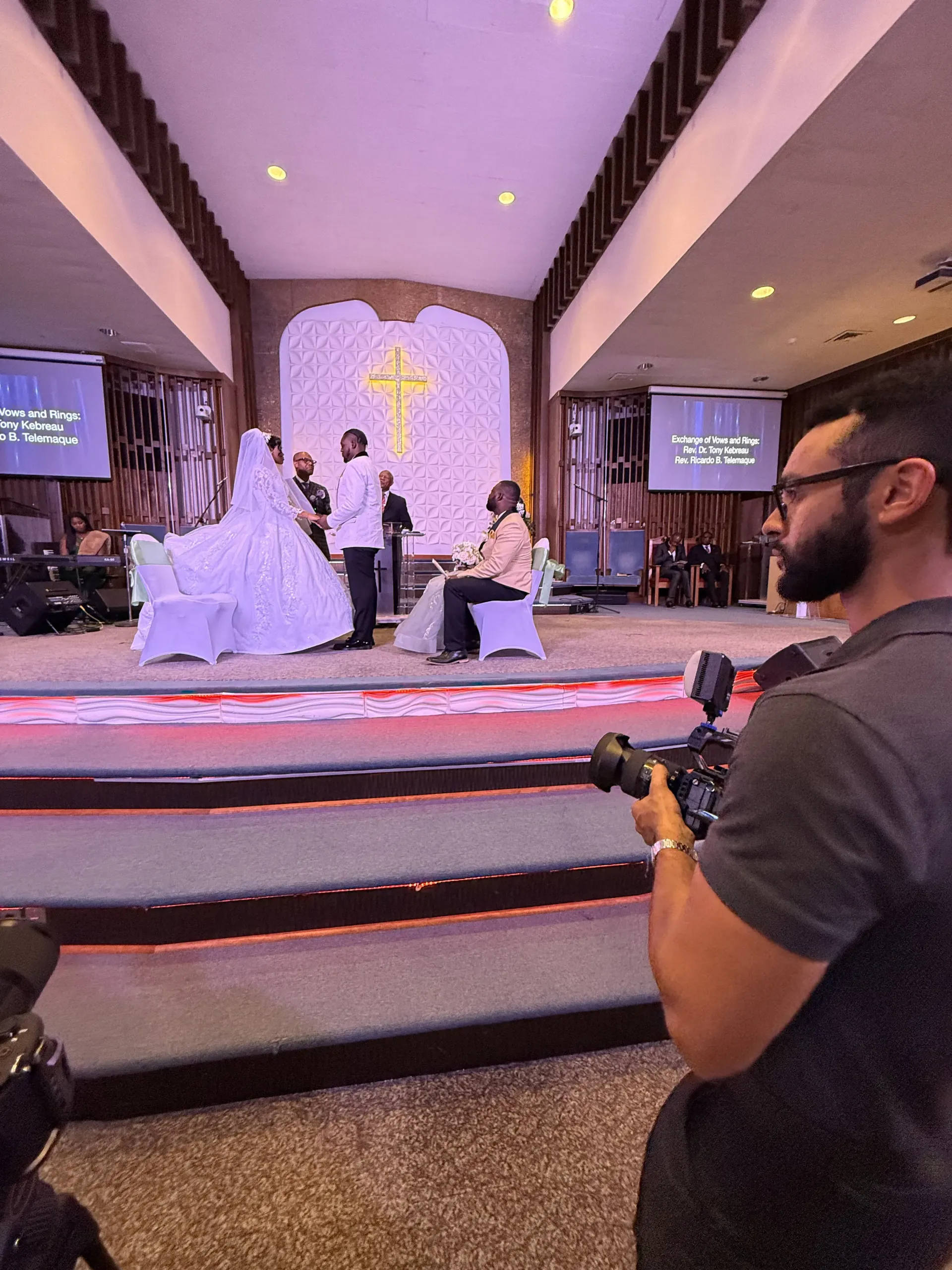 A videographer films a wedding ceremony on a stage with a cross backdrop, while the couple stands before an officiant.