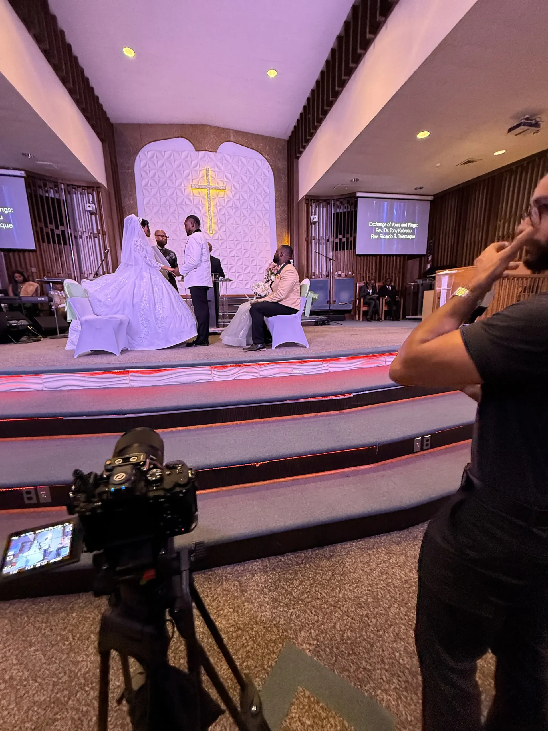 A wedding ceremony taking place on a stage in front of a cross, filmed by a camera in the foreground.