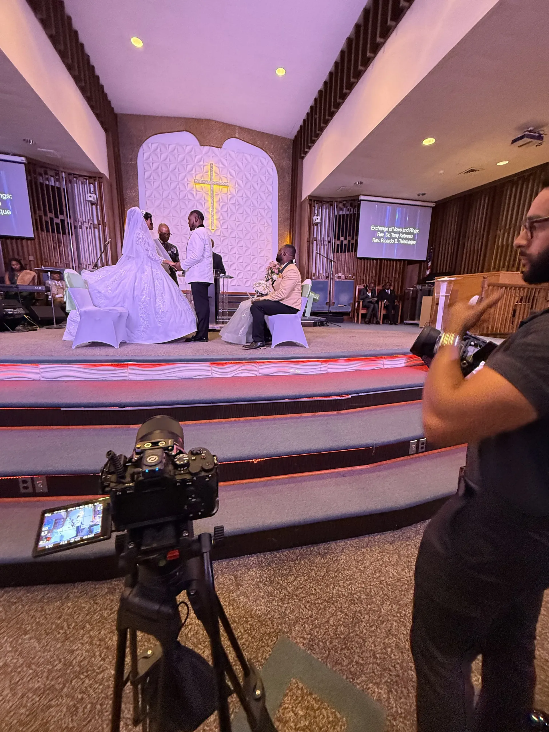 A videographer films a wedding ceremony on a church stage, featuring a couple standing before an officiant.