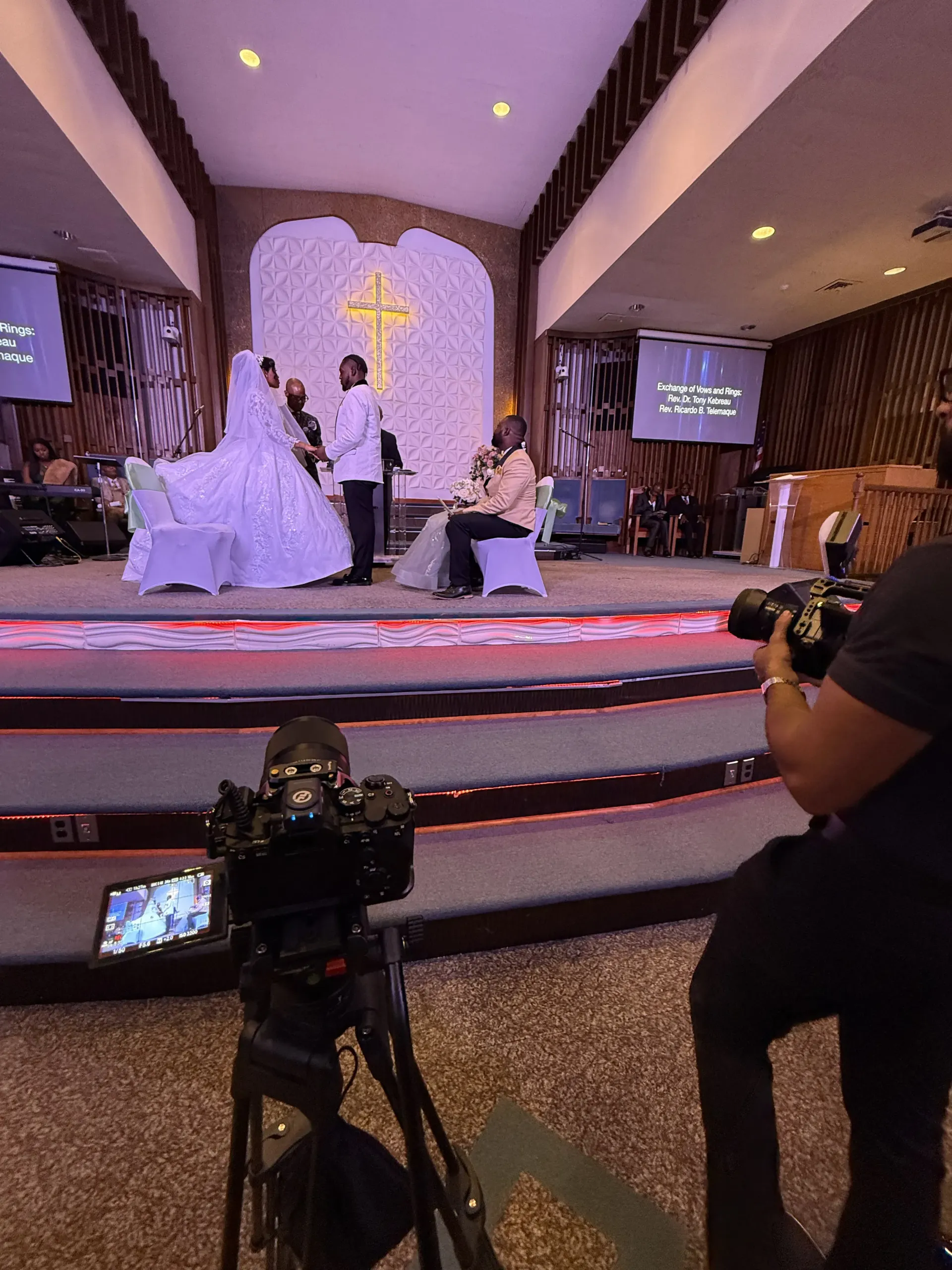 A wedding ceremony taking place on a church stage, filmed by two camera operators in the foreground.