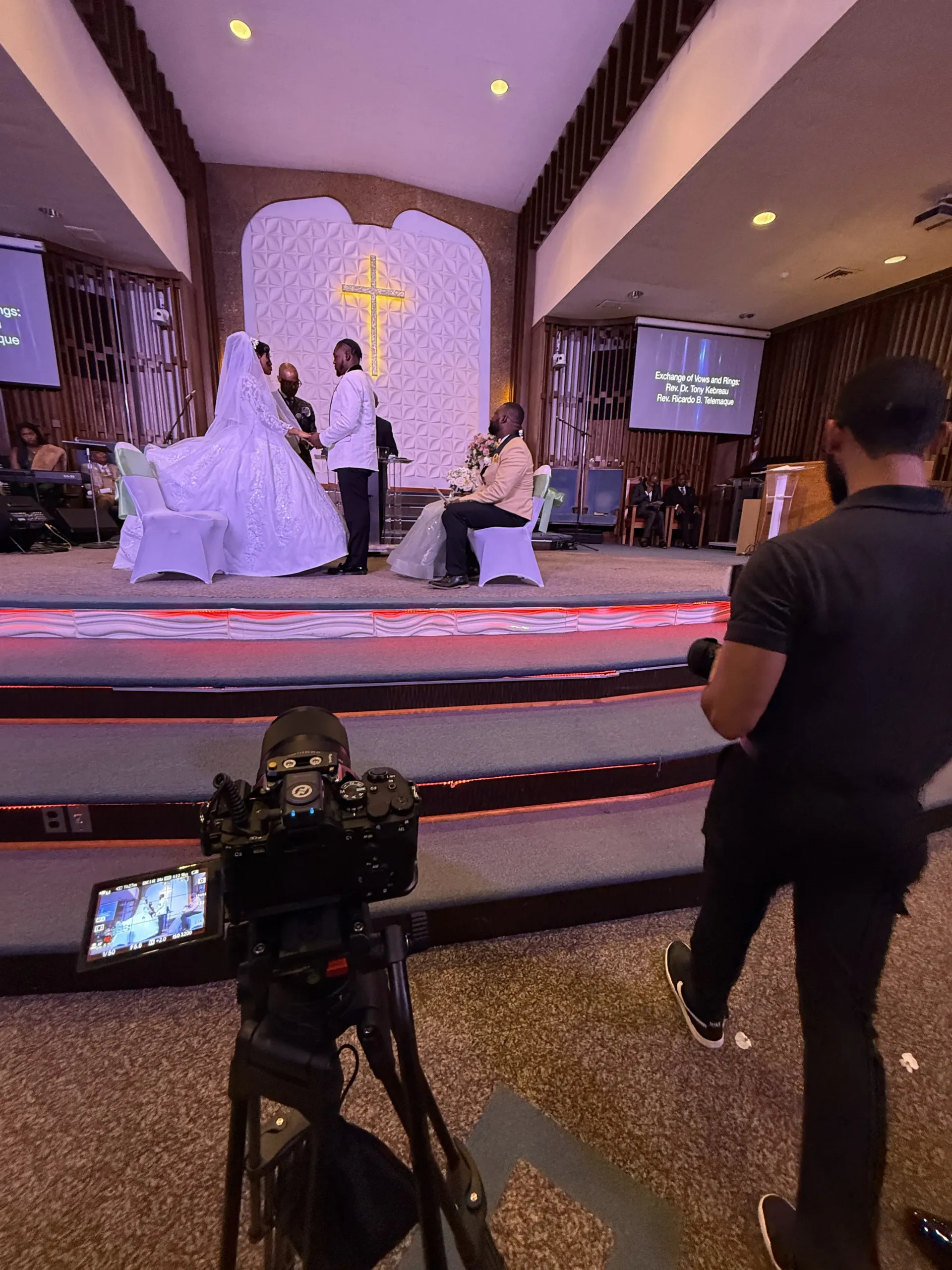 A wedding ceremony in progress, with a bride and groom on stage while a videographer captures the event.