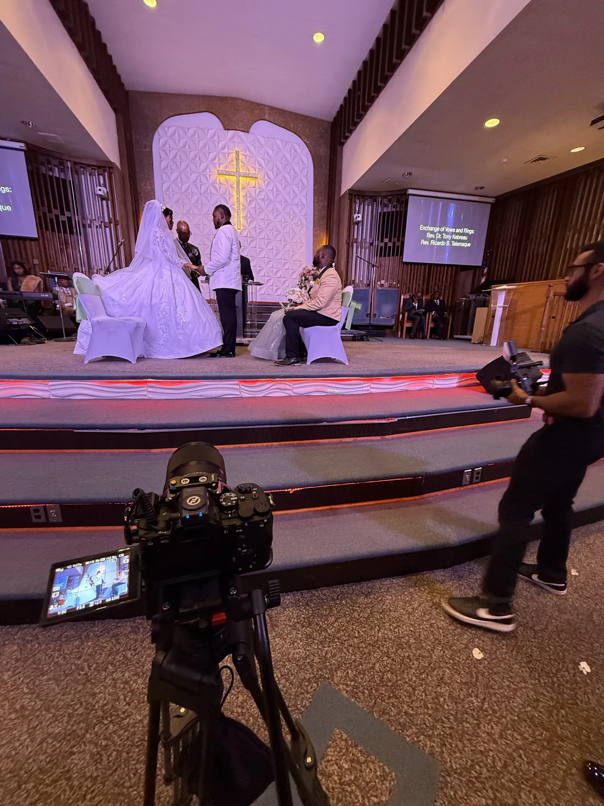 A camera on a tripod films a wedding ceremony taking place on a church stage in front of a large, illuminated cross.