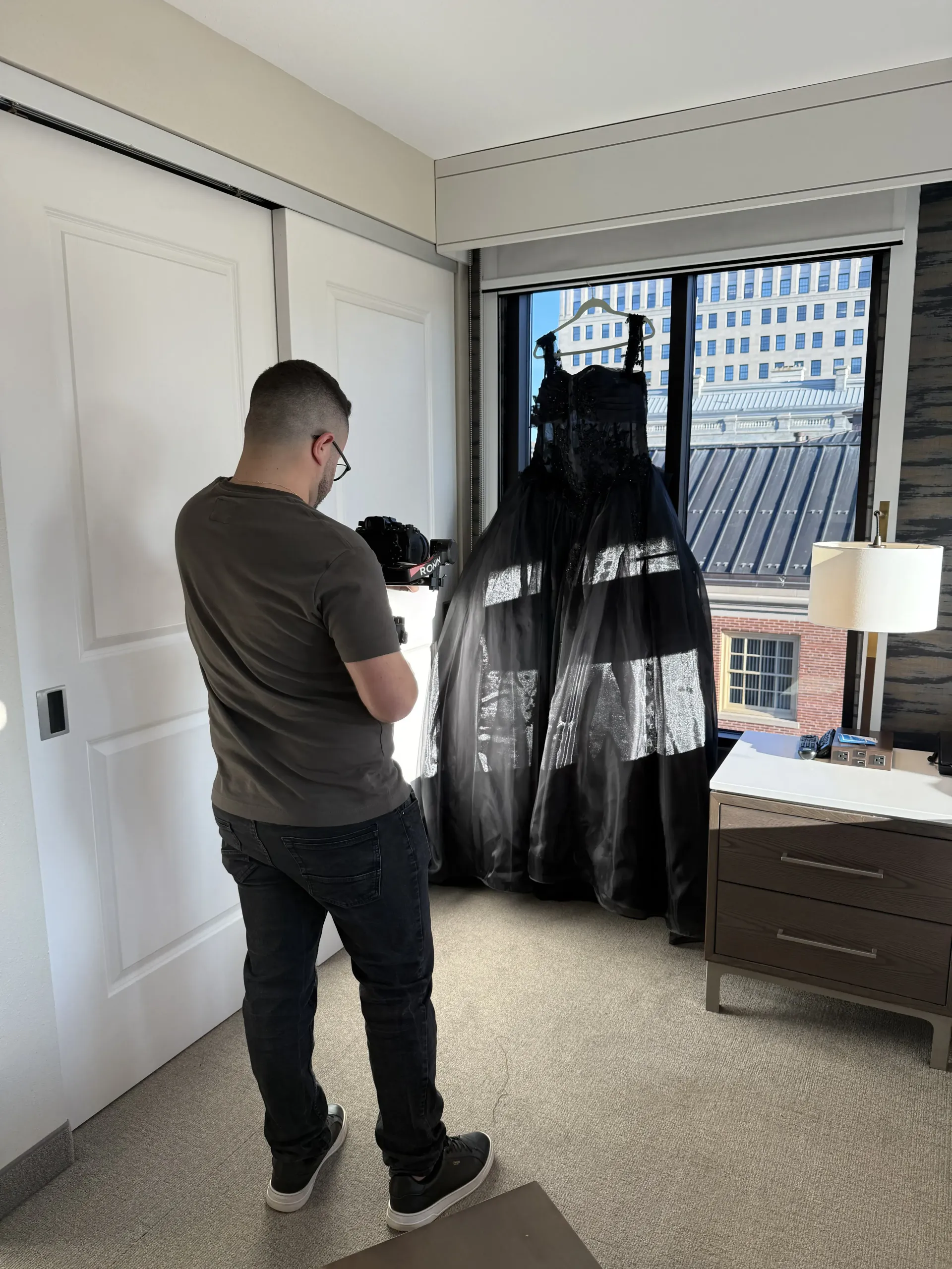 A person in a brown t-shirt and jeans stands in a room, photographing a long, black dress hanging by a window.
