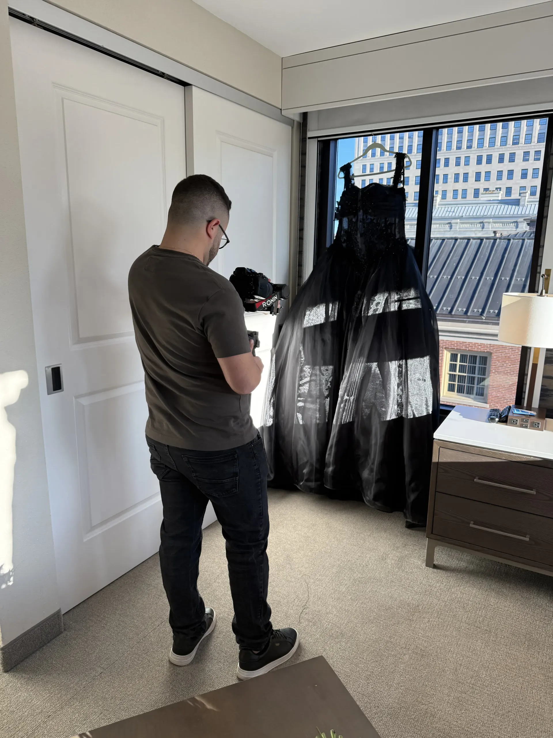 A person in a brown t-shirt and dark jeans uses a camera to photograph a black gown hanging in front of a window.