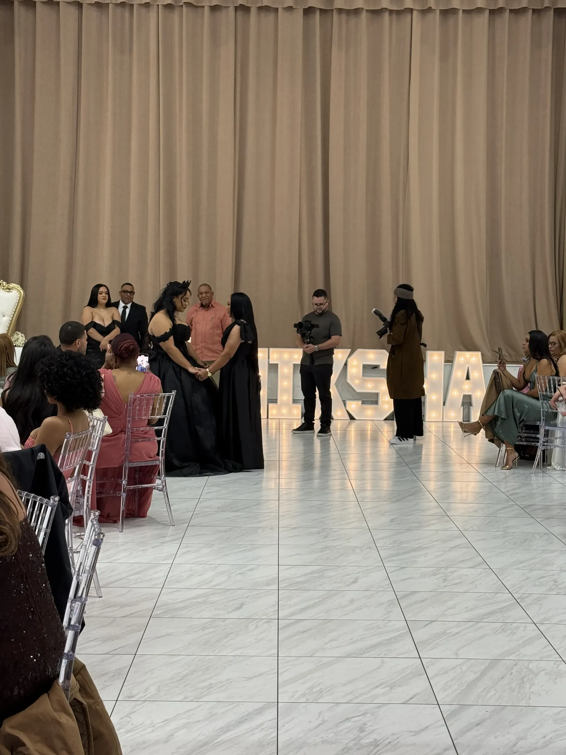 Two people in black gowns hold hands at a formal event, surrounded by guests and large, illuminated letter props.