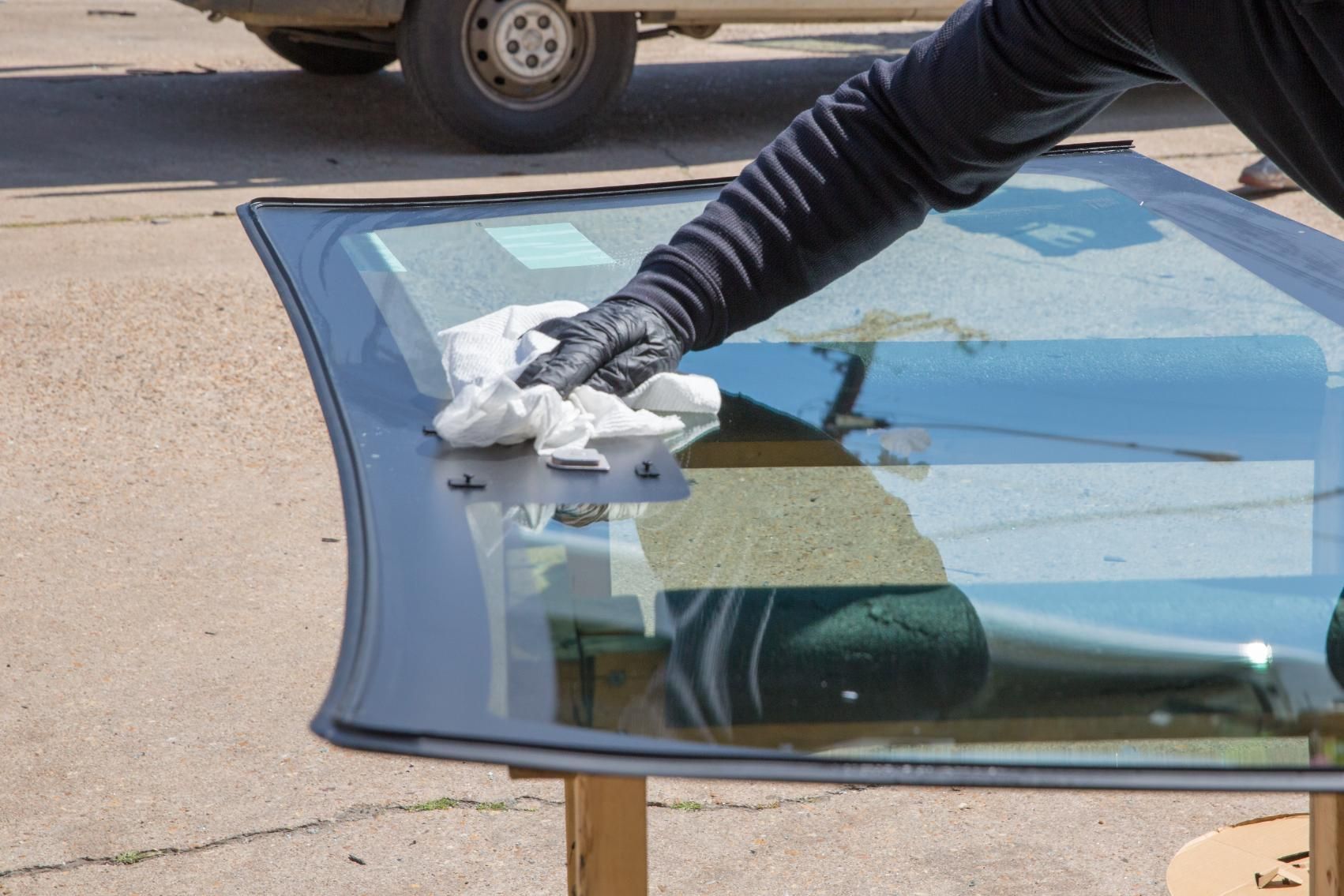 Person in black gloves wiping a car windshield on a table outside, vehicle in background.