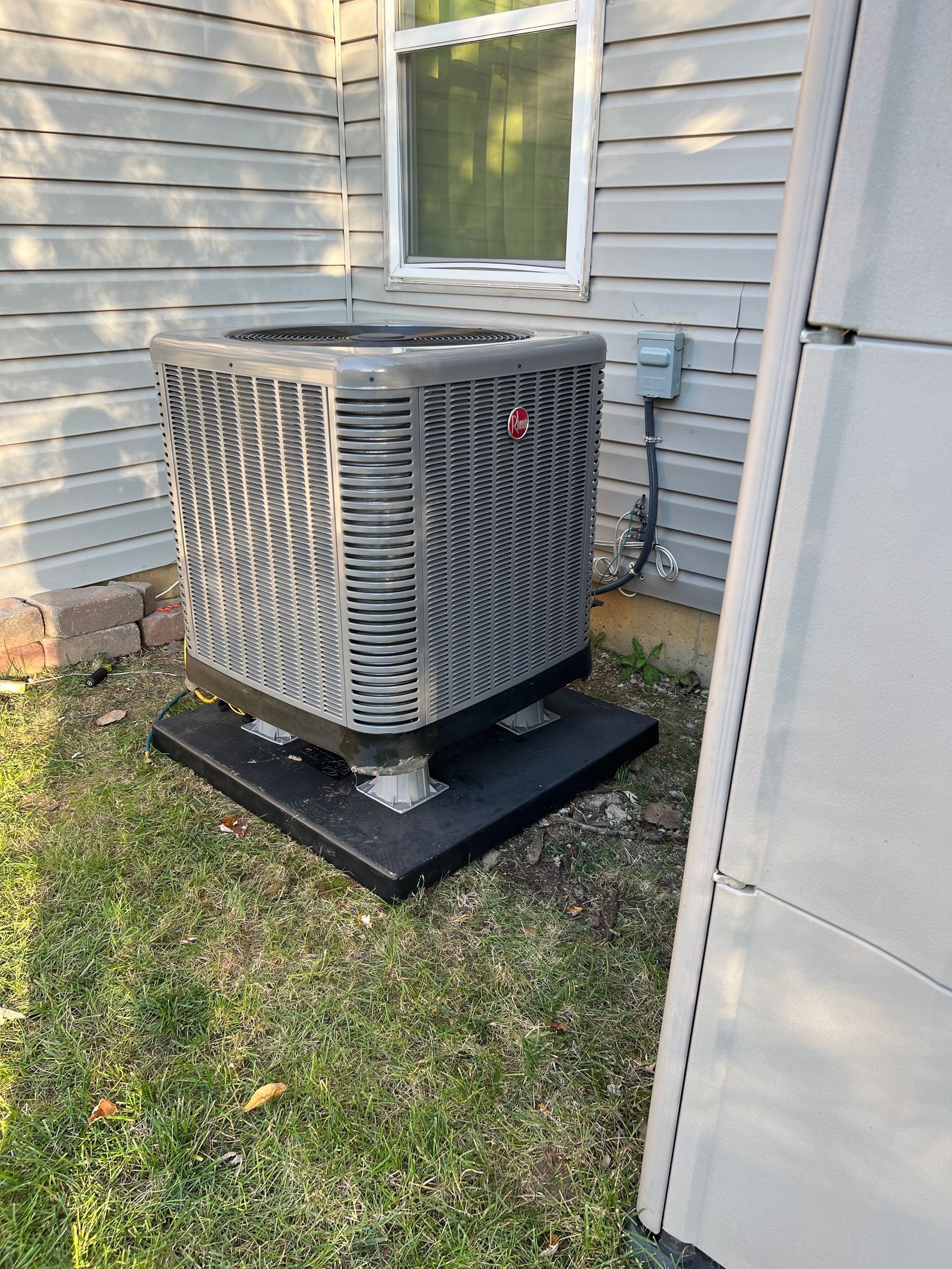 AC unit on black base, beside a building with gray siding and a window.