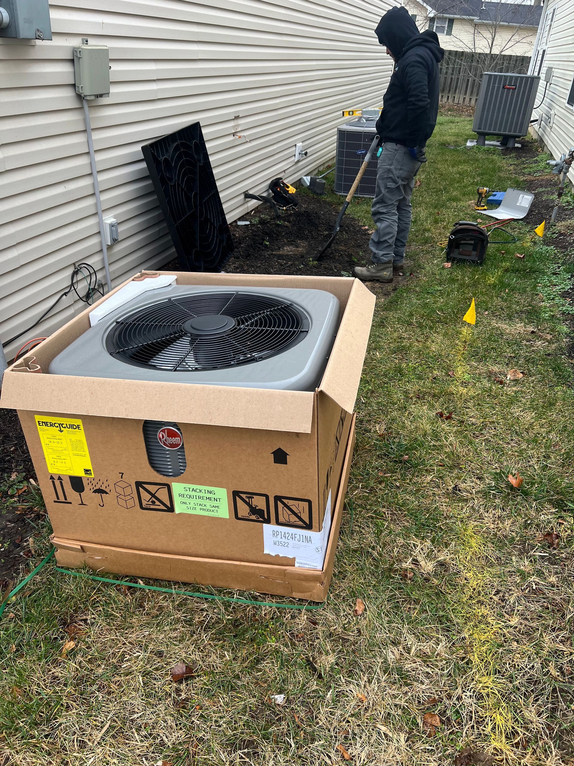 HVAC unit in box, outdoors, with worker nearby. Beige siding and grass visible.