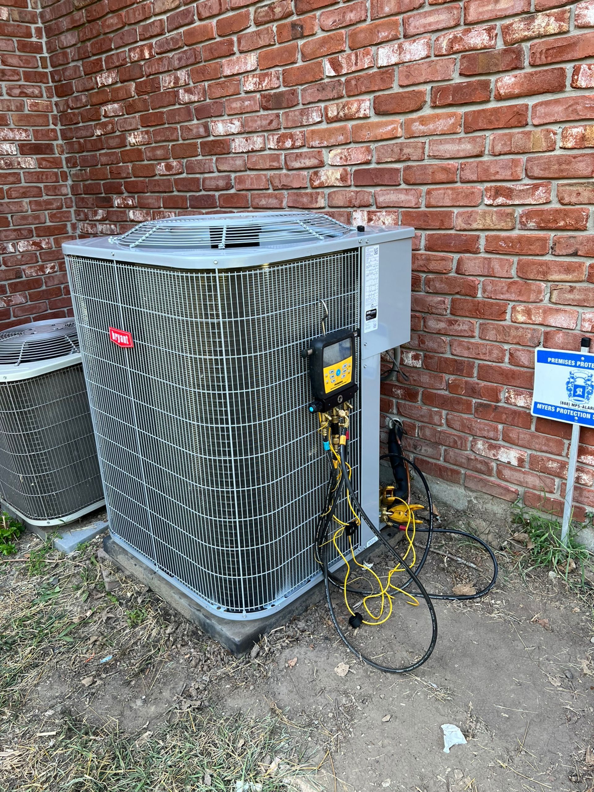 Rheem air conditioning unit with attached equipment next to a brick wall and small blue sign.