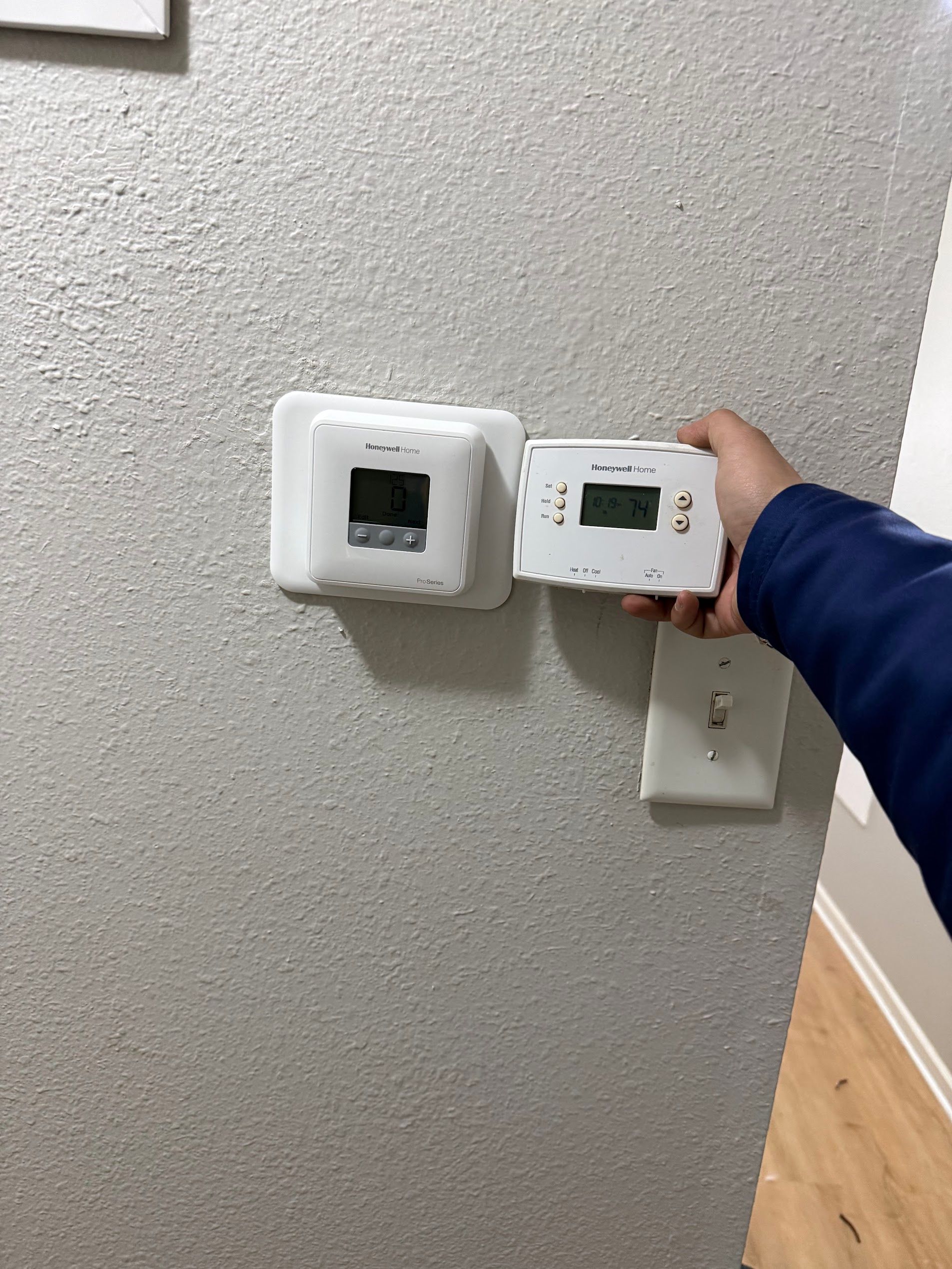 Hand holding a white thermostat on a textured wall beside another wall-mounted control