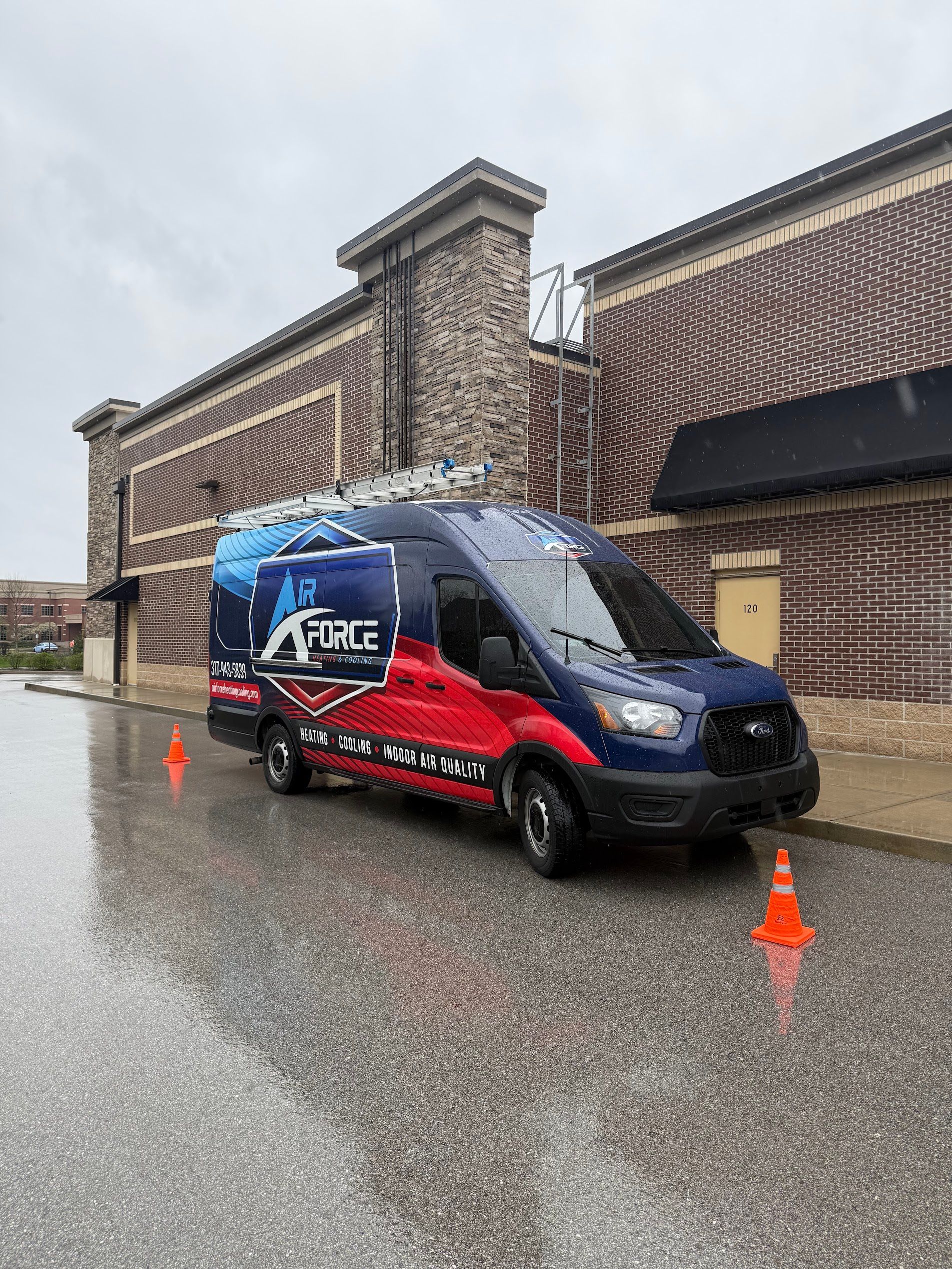 Blue-and-red service van parked by a brick building on a rainy street, with orange cones nearby.