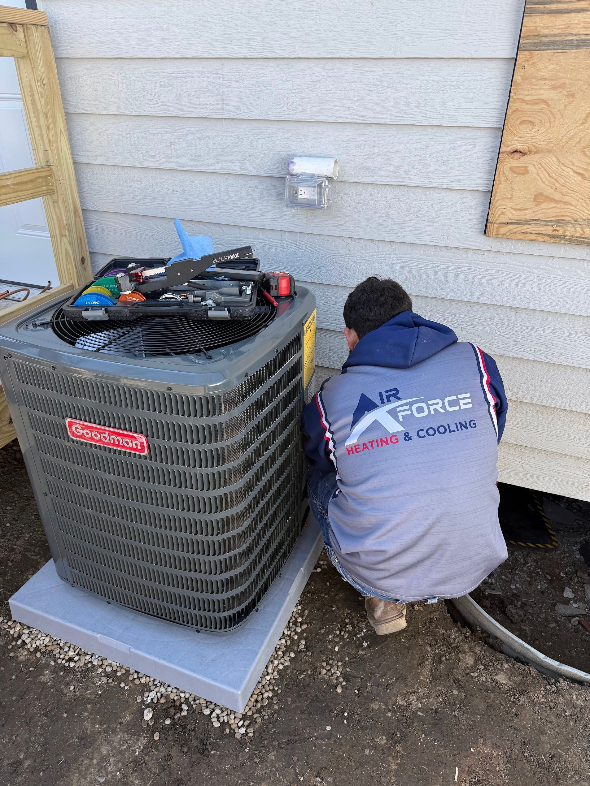 Person kneeling beside an outdoor HVAC unit, working on pipes against a house wall.