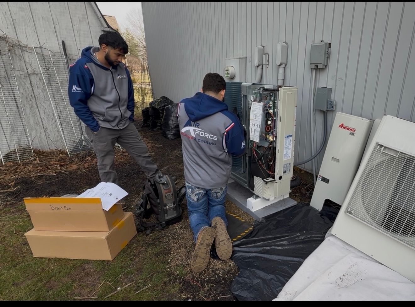 Two technicians inspect outdoor HVAC equipment beside a wall, with tools and boxes on the ground.