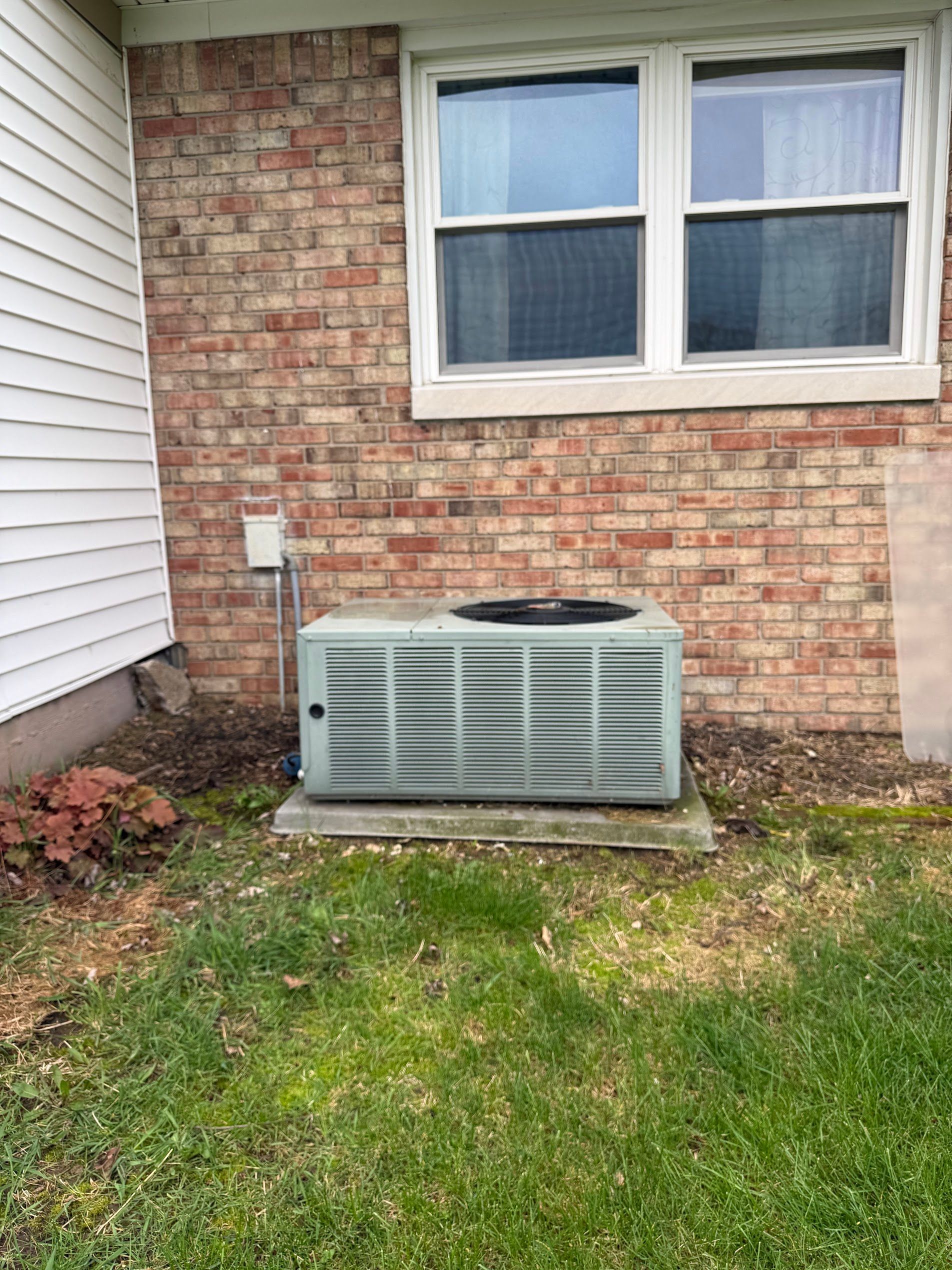 Outdoor HVAC unit beneath a brick house window beside white siding and a small lawn