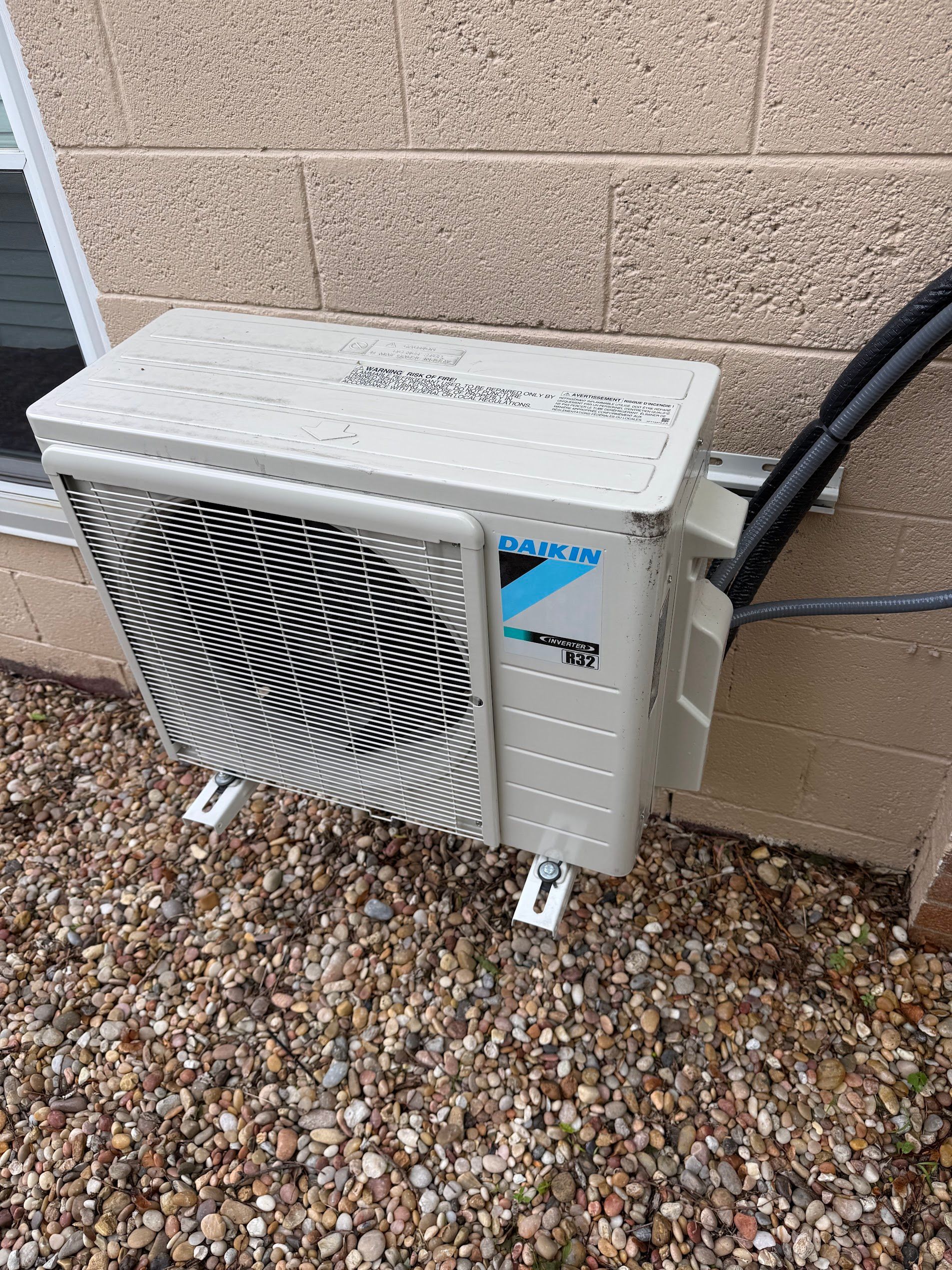 Outdoor wall-mounted air conditioner unit beside a beige brick wall on gravel.