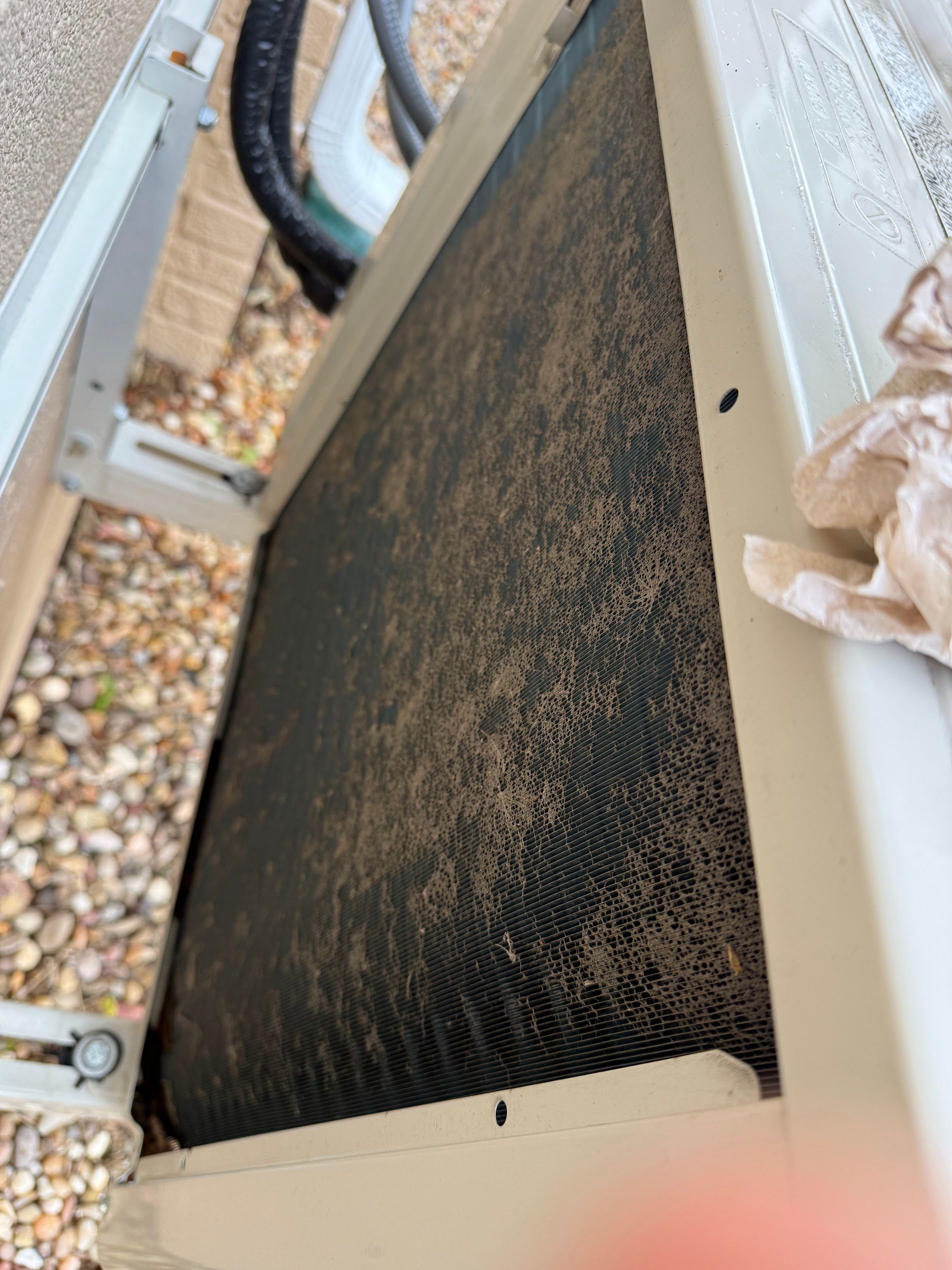 Dirty black HVAC filter in a white outdoor unit, viewed close up among gravel.