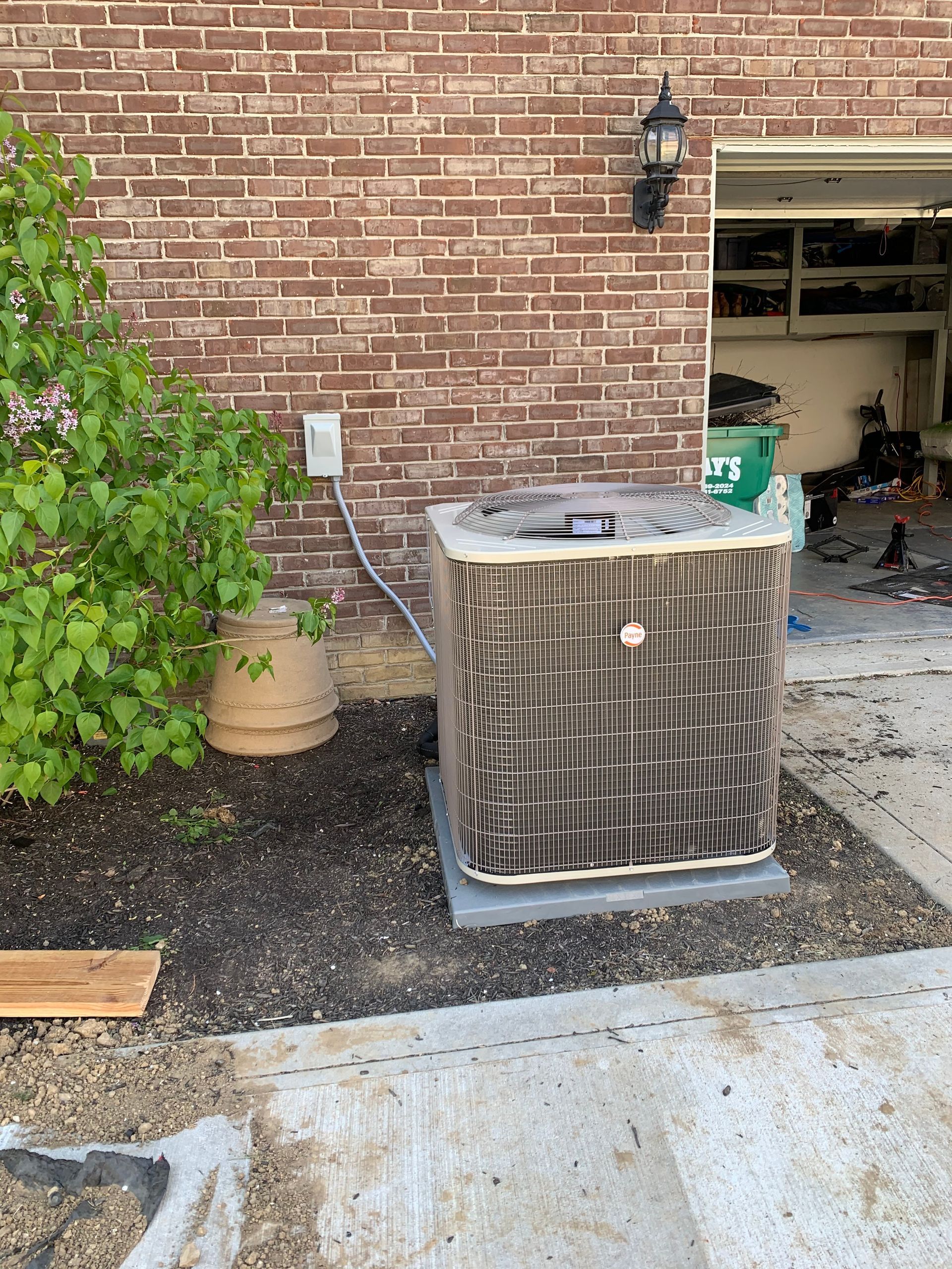 AC unit against brick wall near a garage, next to a bush and decorative pot.