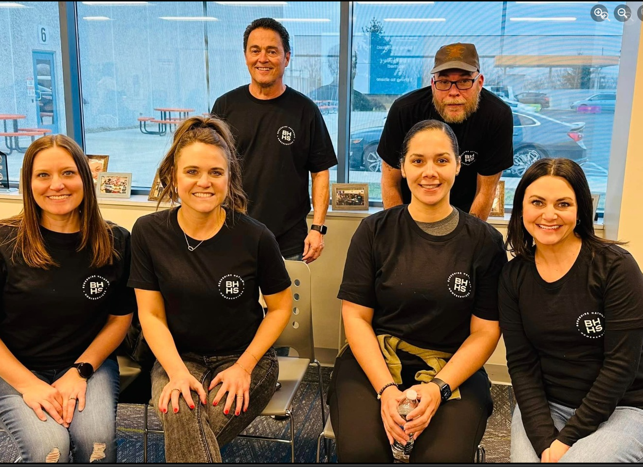Group of six people in black shirts posing indoors. Window backdrop with some vehicles outside.