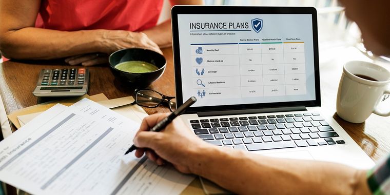 Person writing beside laptop with insurance claims form, calculator, coffee mug, and paperwork on a desk Person writing beside laptop with insurance claims form, calculator, coffee mug, and paperwork on a desk