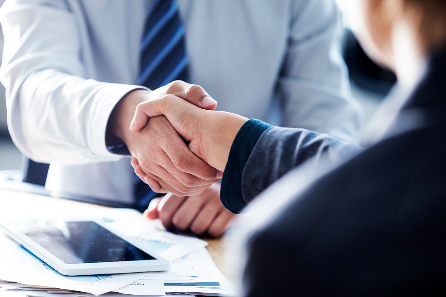 Two businesspeople shaking hands across a desk with a tablet and papers nearby Two businesspeople shaking hands across a desk with a tablet and papers nearby