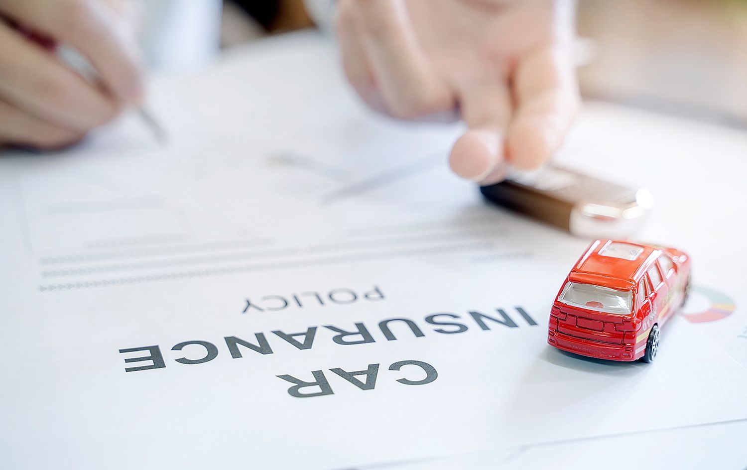 Red toy car on car insurance policy paperwork with a hand and key in the background Red toy car on car insurance policy paperwork with a hand and key in the background