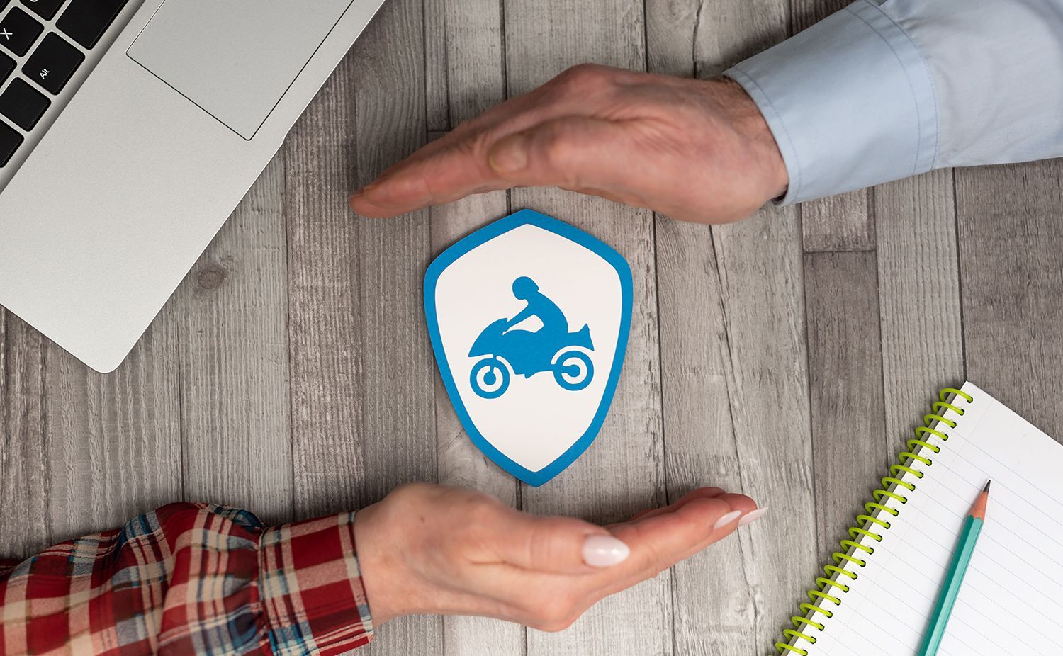 Two hands framing a blue shield icon with a motorcycle on a desk, beside a laptop and notebook. Two hands framing a blue shield icon with a motorcycle on a desk, beside a laptop and notebook.