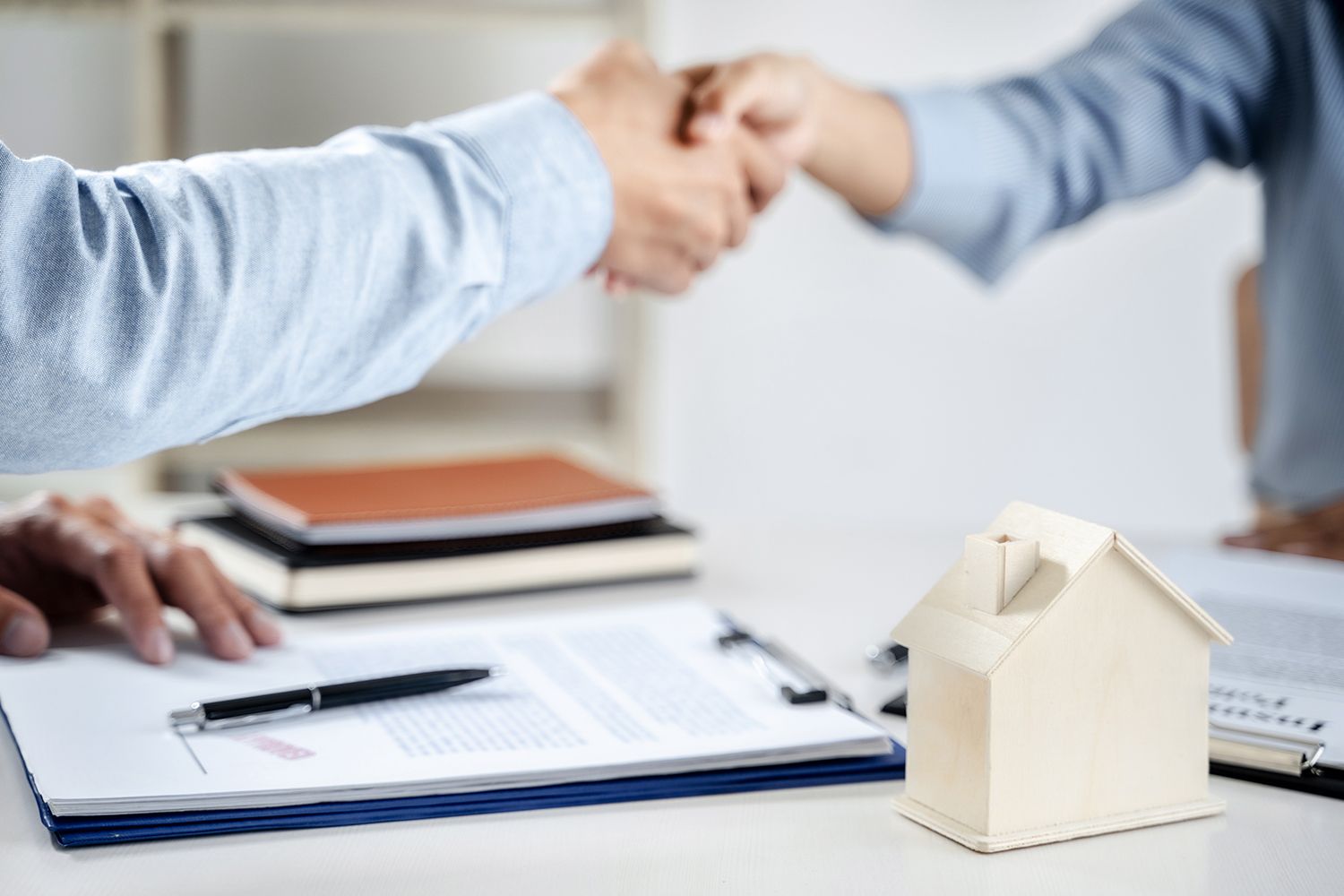 Two people shaking hands over real estate paperwork beside a small house model Two people shaking hands over real estate paperwork beside a small house model