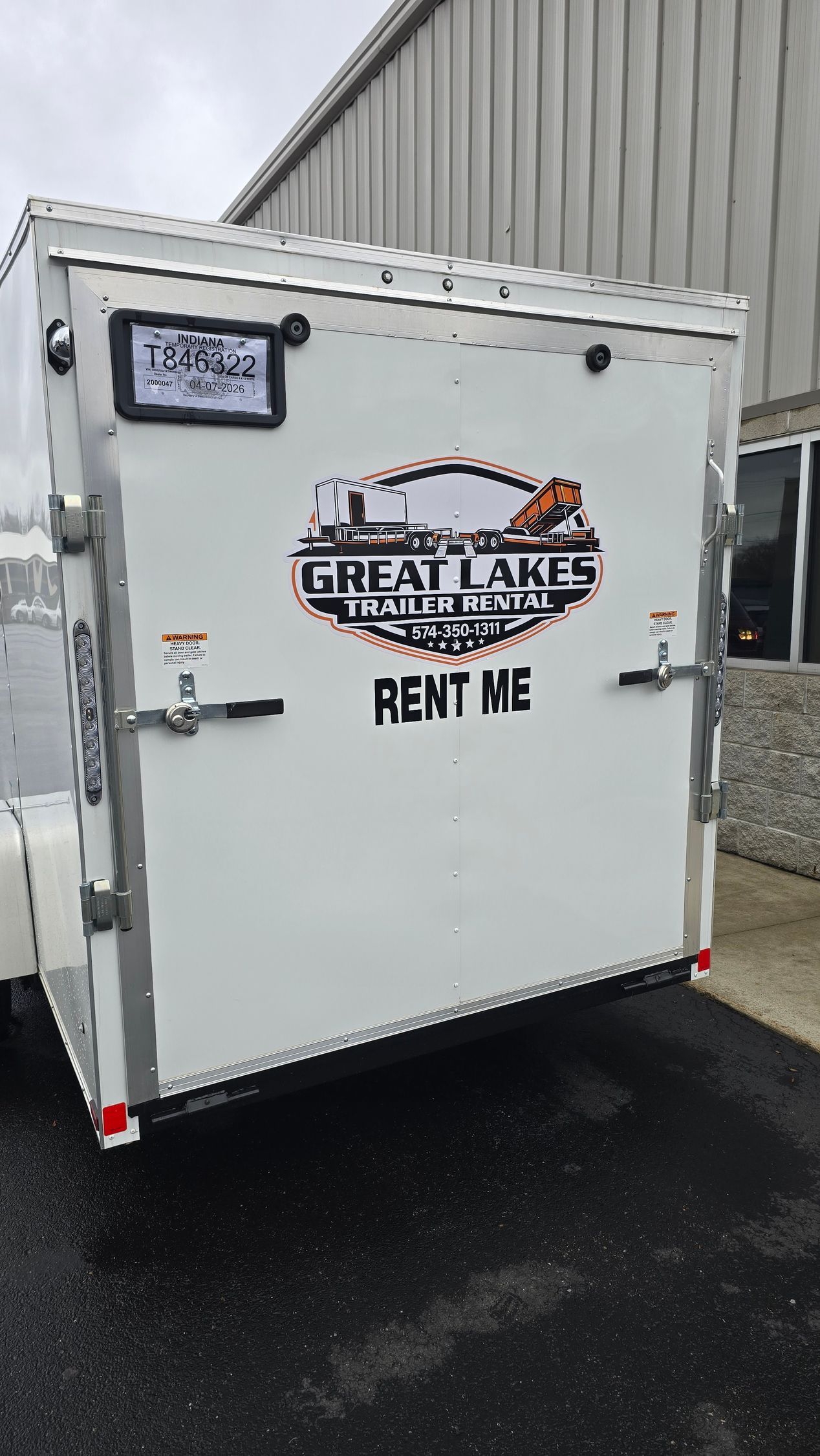 White rental cargo trailer parked on wet pavement beside a gray building.