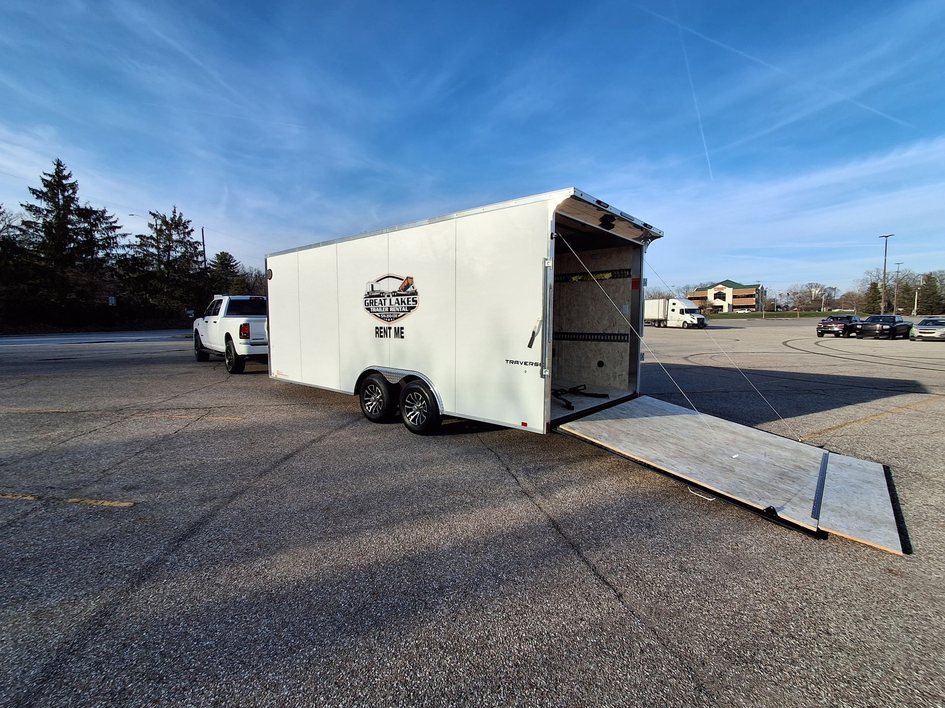 White enclosed trailer with open rear ramp parked on a gravel lot under a blue sky.