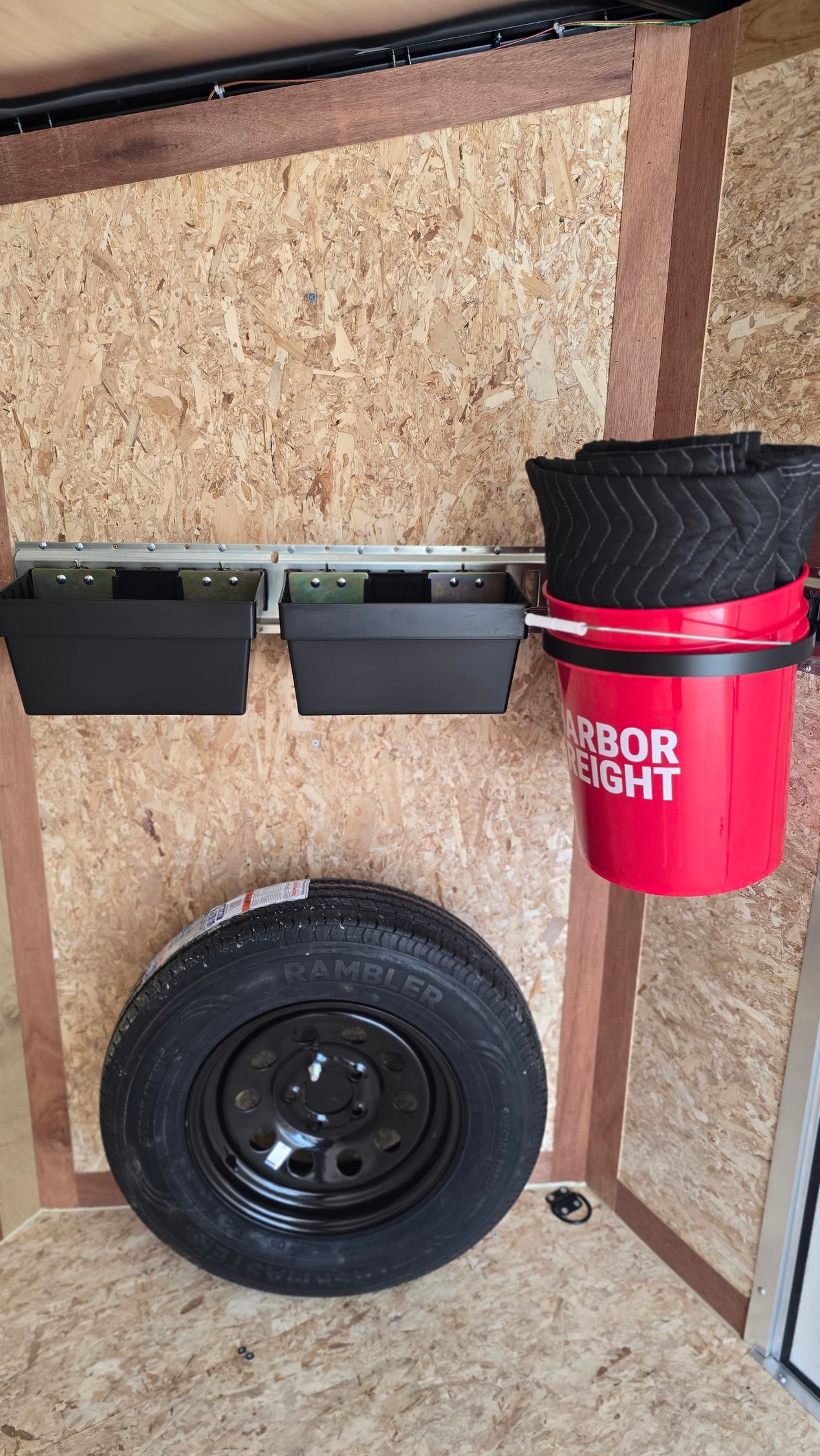Red bucket with lid and two black wall bins above a spare tire mounted on a corkboard wall.