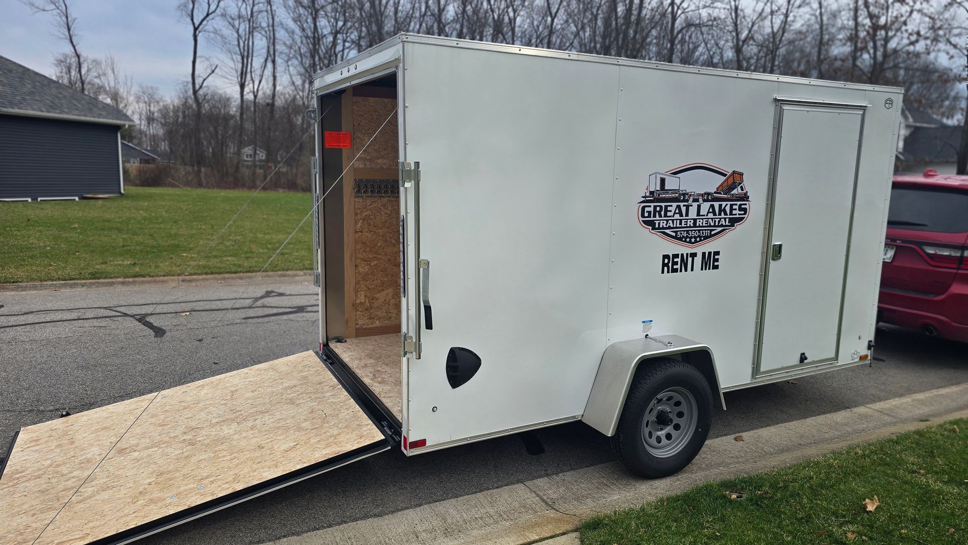 White cargo trailer with open rear ramp parked on a driveway beside a grassy yard and road.
