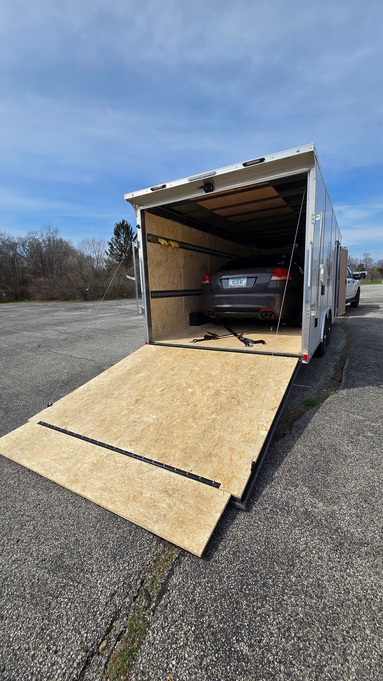 Open car trailer with rear ramp down, showing a car loaded inside on a gravel lot.