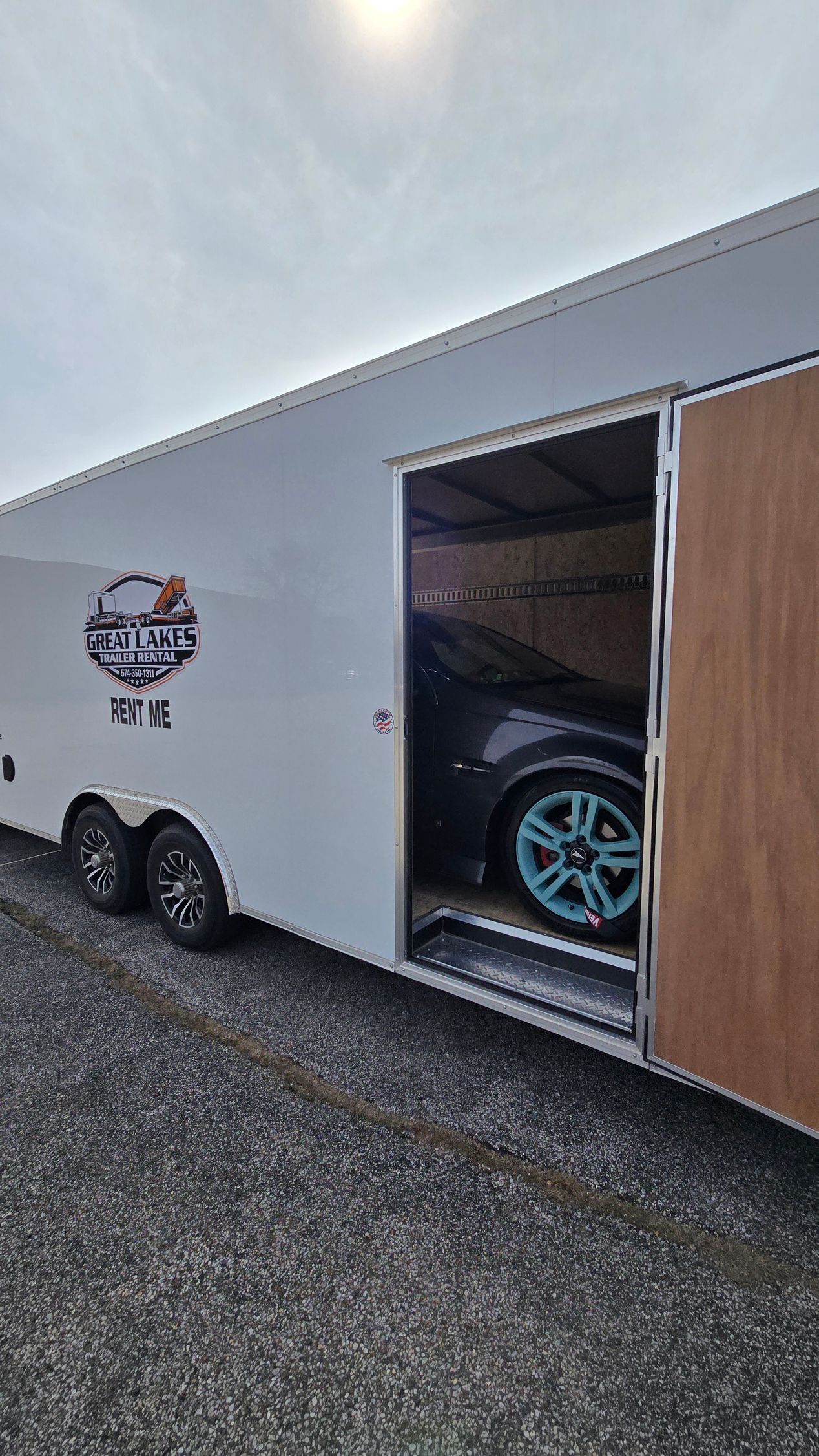 White enclosed trailer with an open side door revealing a car inside on a gravel lot.