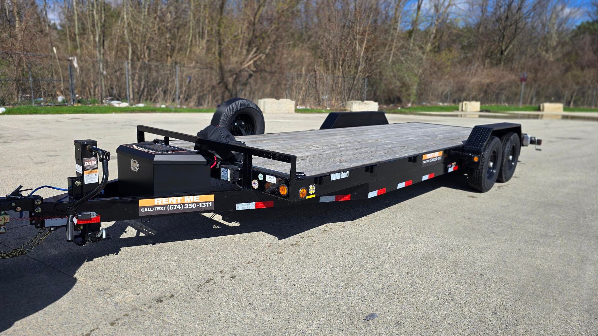 Black flatbed utility trailer parked on gravel with spare tire and toolbox, trees in the background