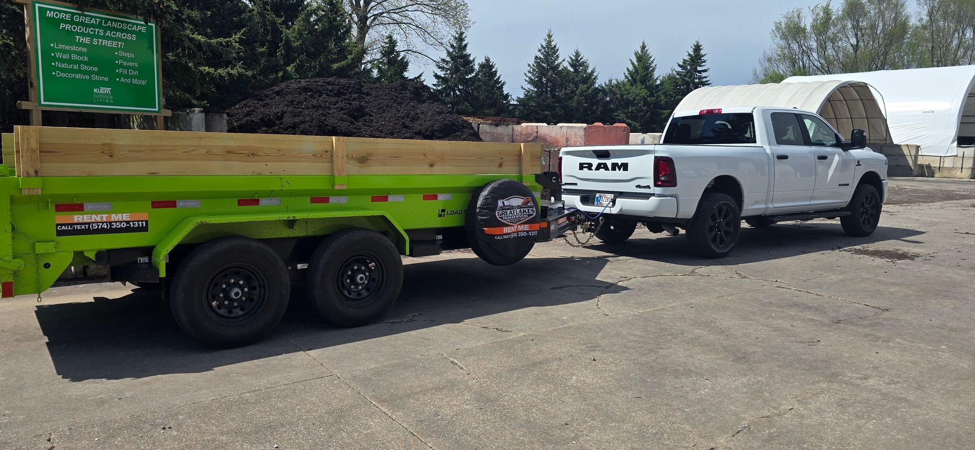 White pickup truck towing a green utility trailer in a dirt lot, with trees in the background