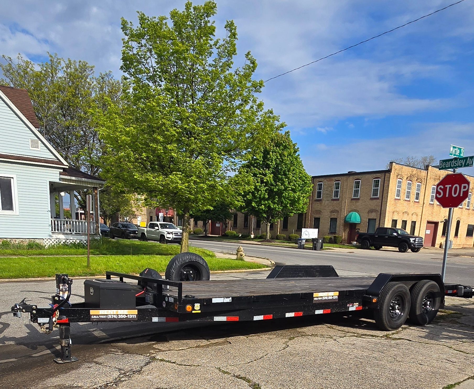Black flatbed trailer parked on a street near a stop sign and brick buildings on a sunny day in Goshen Indiana.