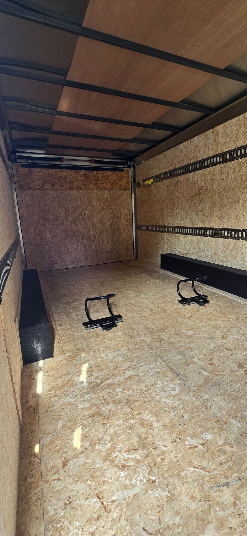 Empty cargo trailer interior with wooden walls, black roof supports, and two tie-down straps on the floor
