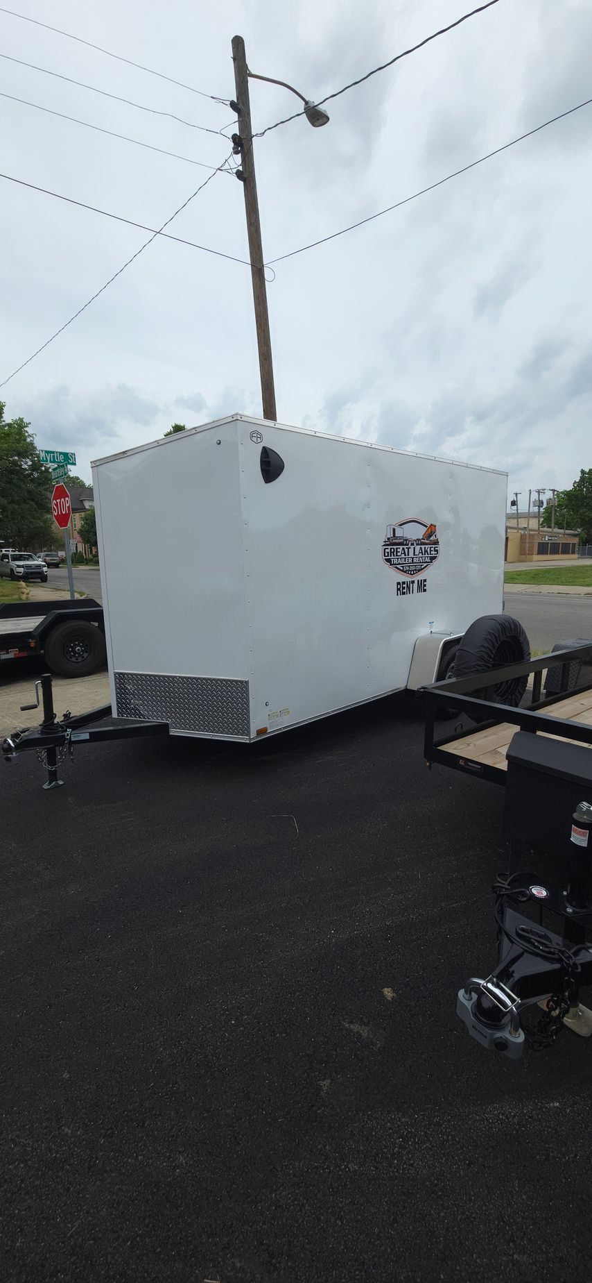 White enclosed trailer parked on wet pavement under cloudy skies, with utility poles nearby.