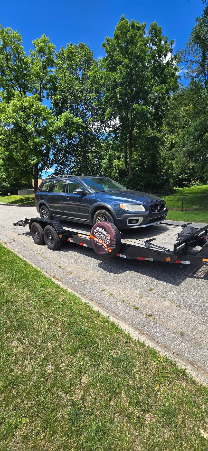 Black SUV on a flatbed trailer parked on a gravel driveway beside green trees under a blue sky