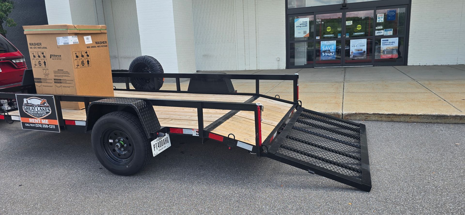 Small black trailer with folded metal ramp parked outside a storefront, carrying a cardboard box.