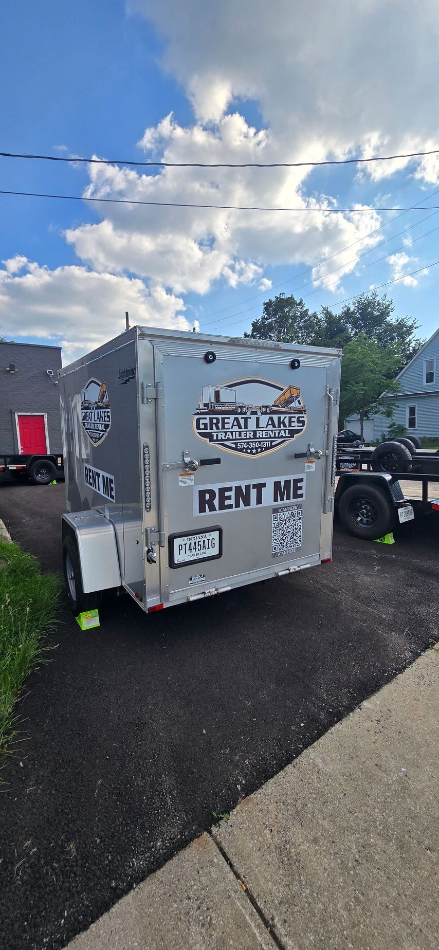 Gray RENT ME trailer in a parking lot under a partly cloudy sky, with a truck beside it.