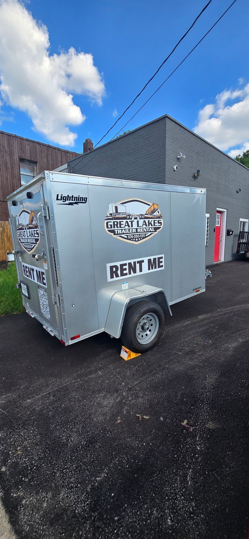 White rental utility trailer with “RENT ME” sign parked on an asphalt lot beside buildings.