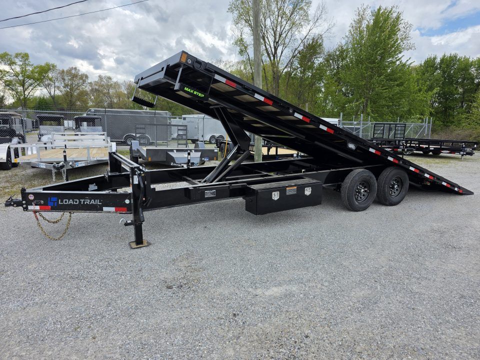 Black dual-axle car hauler trailer with ramp raised in a gravel lot, trees and trucks in background