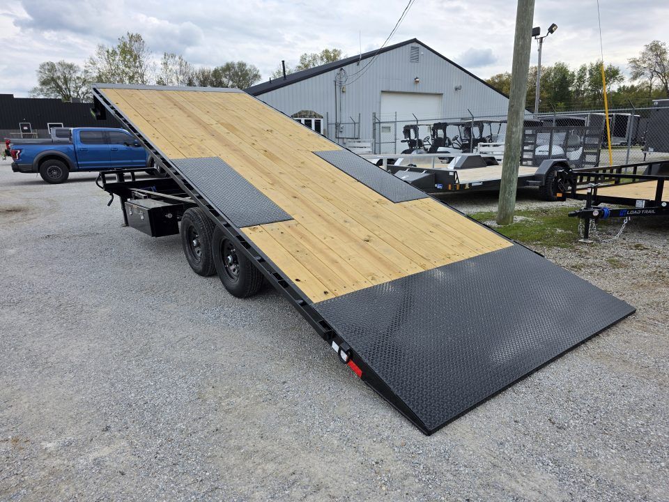 Tilted black-and-wood trailer ramp in a gravel lot, with pickup trucks and a warehouse in the background