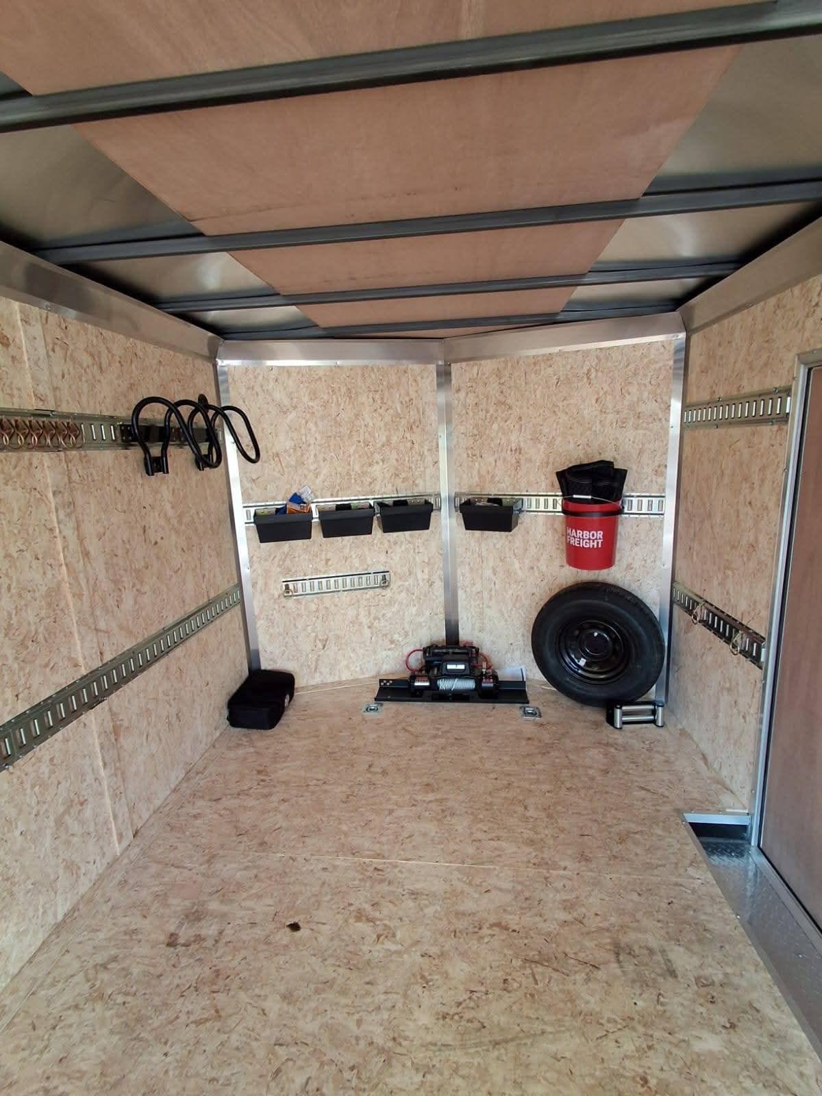 Interior of an empty cargo trailer with tools, a spare tire, and a red bucket along the walls.