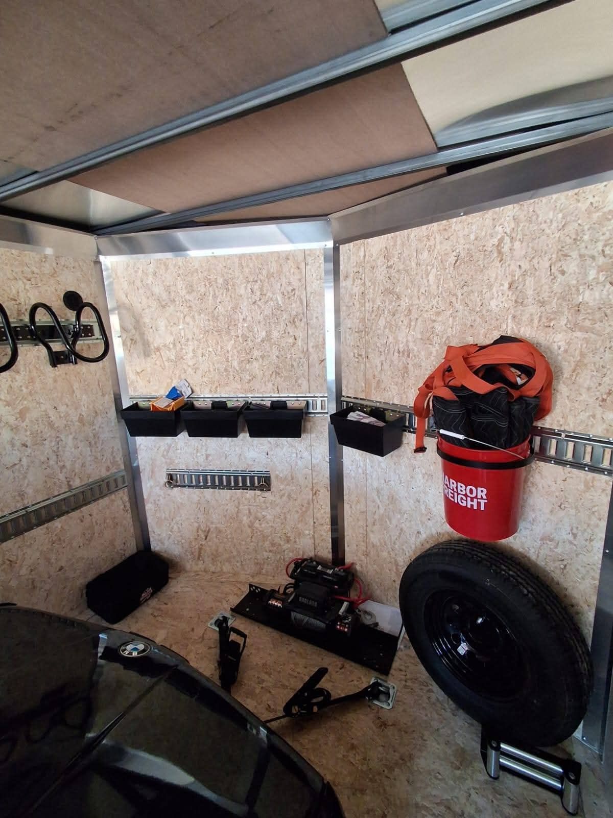 Small trailer interior with tools, a spare tire, and red bucket against plywood walls.