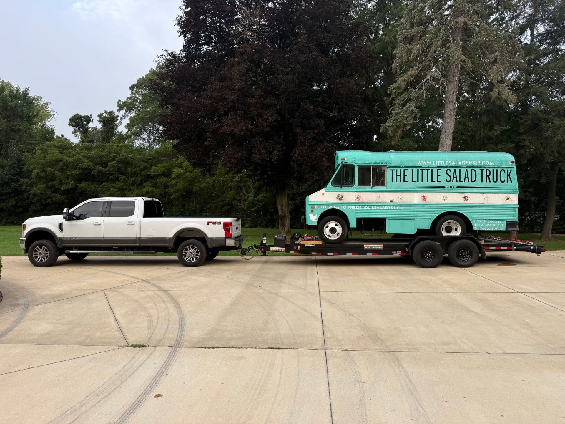 White pickup towing a turquoise mobile sauna trailer on a road near trees